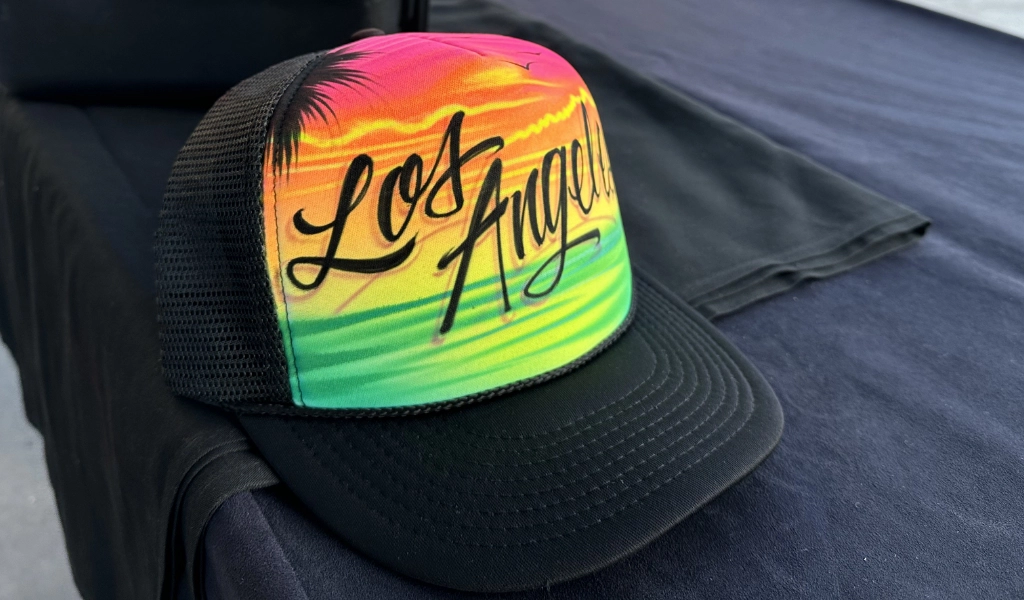 A trucker hat with a vibrant, airbrush art-inspired sunset design and "Los Angeles" proudly displayed on the front, resting on a dark surface.