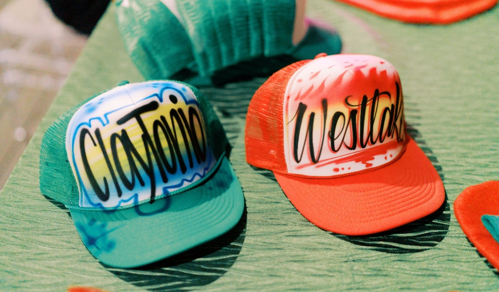 Two trucker hats sit on a table, showcasing vibrant airbrush art. One boldly displays "Clayton" in blue and green, while the other features "Westlake" in striking red and white.
