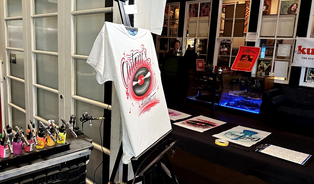 A white T-shirt featuring a vinyl record graphic is showcased on an easel, surrounded by airbrushed designs and paint supplies. Posters adorn the room's walls, capturing the dynamic essence of live art favored by airbrush artists for events.