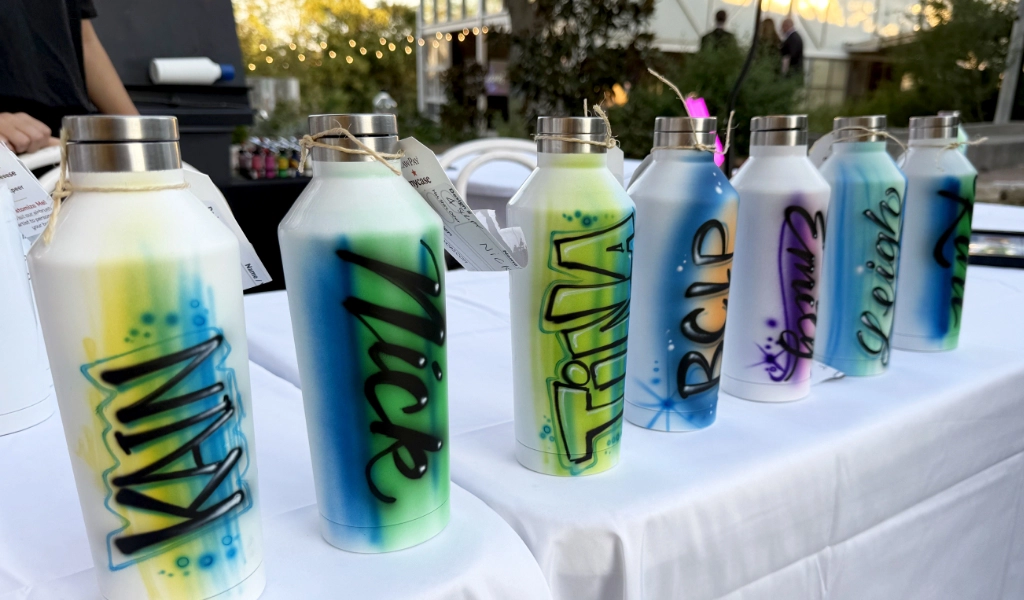 Six personalized, airbrushed bottles with names displayed on a table, each featuring vibrant colors and unique styles.