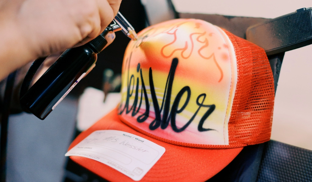 A person airbrushes a red and yellow trucker hat with graffiti-style text. A name tag is attached to the hat.