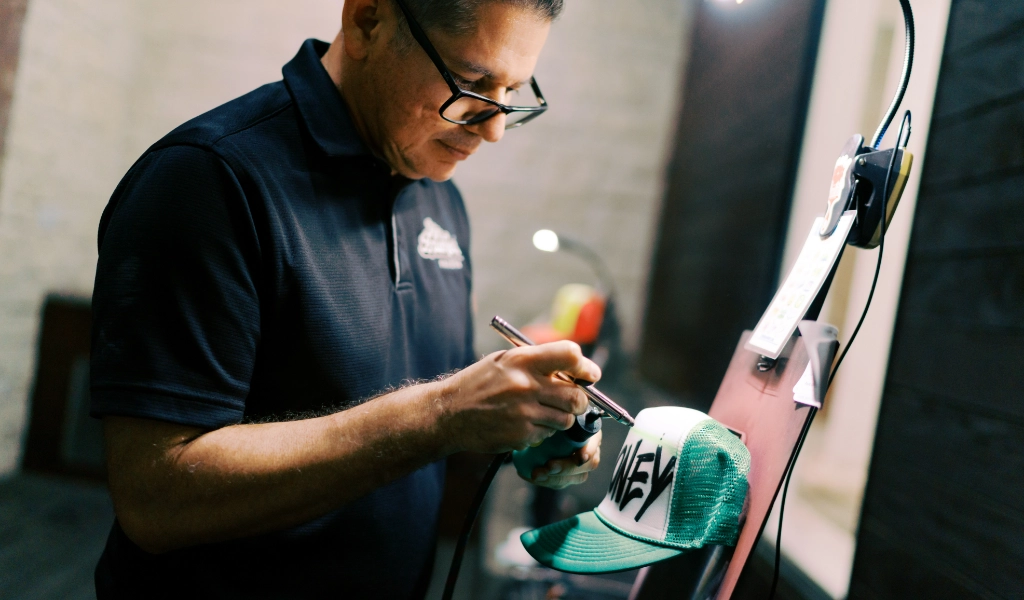 A live airbrush artist skillfully paints a green and white trucker hat, adorning it with a word in bold black letters, bringing the design to life with vibrant airbrush art.