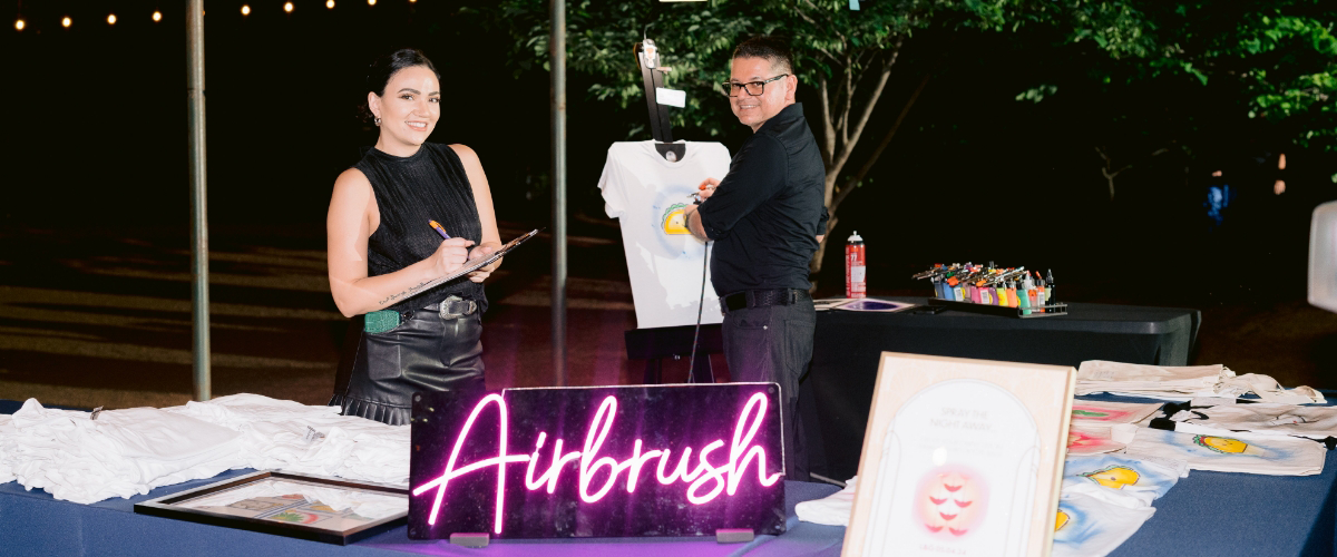 Two people at a booth with a neon "Airbrush" sign; one is smiling with a clipboard, the other showcasing live art by airbrushing a T-shirt.
